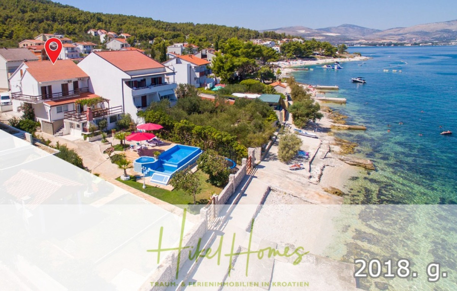 1. Reihe dirkt Meer mit 5 SZ, Pool, Jacuzzi, … auf der Insel Ciovo (bei Trogir mit 2 Brücken) - Luftaufnahme von Häusern am Meer mit einem Pool, Gärten und einer felsigen Küste in Kroatien. Ein roter Standortmarker hebt ein Haus hervor. Blaues Meer und grüne Hügel im Hintergrund. Die Textüberlagerung lautet "Hikel Homes" und "2018. g.". (44915)