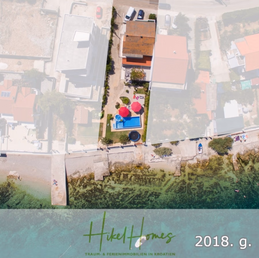 1. Reihe dirkt Meer mit 5 SZ, Pool, Jacuzzi, … auf der Insel Ciovo (bei Trogir mit 2 Brücken) - Luftaufnahme eines Küstengrundstücks mit Haus, Pool und Bäumen. Das Grundstück liegt direkt am Strand. Das Bild ist mit Branding und Text in einer Ecke überlagert, was auf Immobilien in Kroatien hindeutet, mit der Angabe des Jahres 2018. (42951)