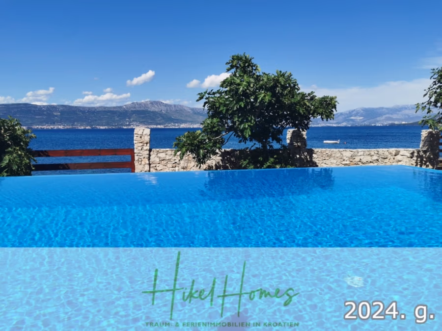 1. Reihe dirkt Meer mit 5 SZ, Pool, Jacuzzi, … auf der Insel Ciovo (bei Trogir mit 2 Brücken) - Infinity-Pool mit Blick auf das ruhige Meer unter klarem, blauem Himmel, in der Ferne mit Bergen und vereinzelten Wolken. Steinmauer und Bäume trennen den Pool vom Meer. Unten steht „Hikel Homes“ und „2024. g.“. (42955)