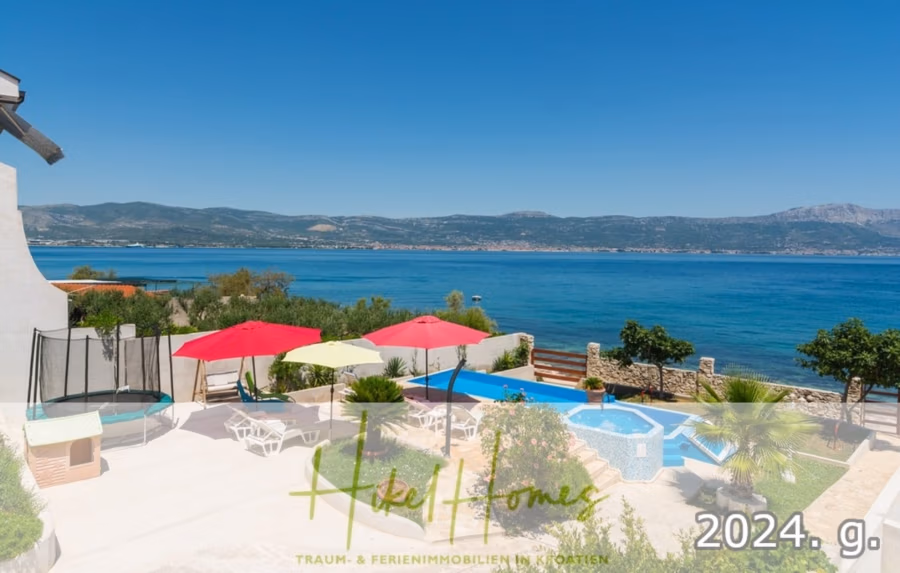 1. Reihe dirkt Meer mit 5 SZ, Pool, Jacuzzi, … auf der Insel Ciovo (bei Trogir mit 2 Brücken) - Ein Küstenblick mit Pool und Terrasse, roten und rosa Sonnenschirmen, Liegestühlen und umgebendem Grün. Im Hintergrund das blaue Meer und die fernen Hügel. Textüberlagerungen lauten „2024“ und „Hotel Homes“. (42923)