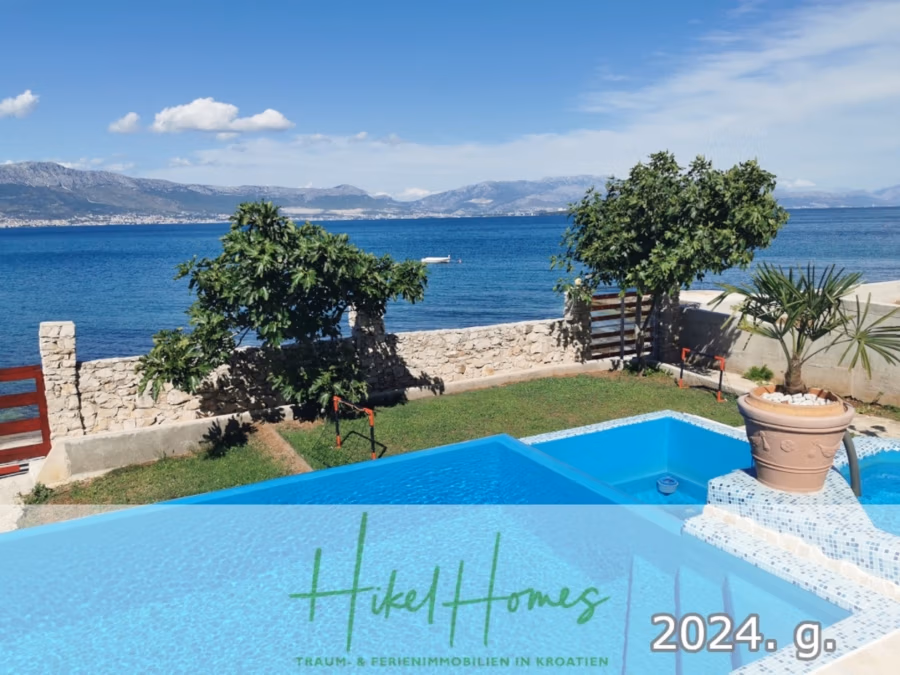 1. Reihe dirkt Meer mit 5 SZ, Pool, Jacuzzi, … auf der Insel Ciovo (bei Trogir mit 2 Brücken) - Ein malerischer Blick auf eine Küstenlandschaft mit Swimmingpool, üppigem Grün und einer Steinmauer mit Blick auf das Meer. In der Ferne sind Berge unter blauem Himmel zu sehen. Textüberlagerungen lauten: „Hikel Homes“ und „2024. g.“ auf Deutsch und Kroatisch. (42953)