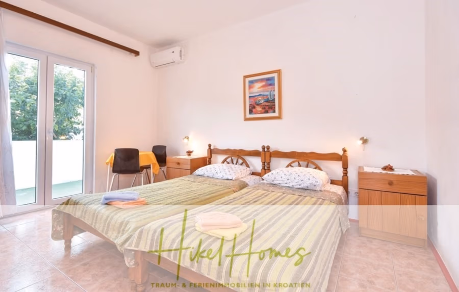 1. Reihe dirkt Meer mit 5 SZ, Pool, Jacuzzi, … auf der Insel Ciovo (bei Trogir mit 2 Brücken) - Ein helles Schlafzimmer mit zwei zusammengeschobenen Einzelbetten und gestreiften grün-gelben Decken. Durch Glastüren links gelangt man auf einen kleinen Balkon. Über den Betten hängt ein gerahmtes Bild an der weißen Wand, in der Ecke steht ein schwarzer Stuhl. (42933)