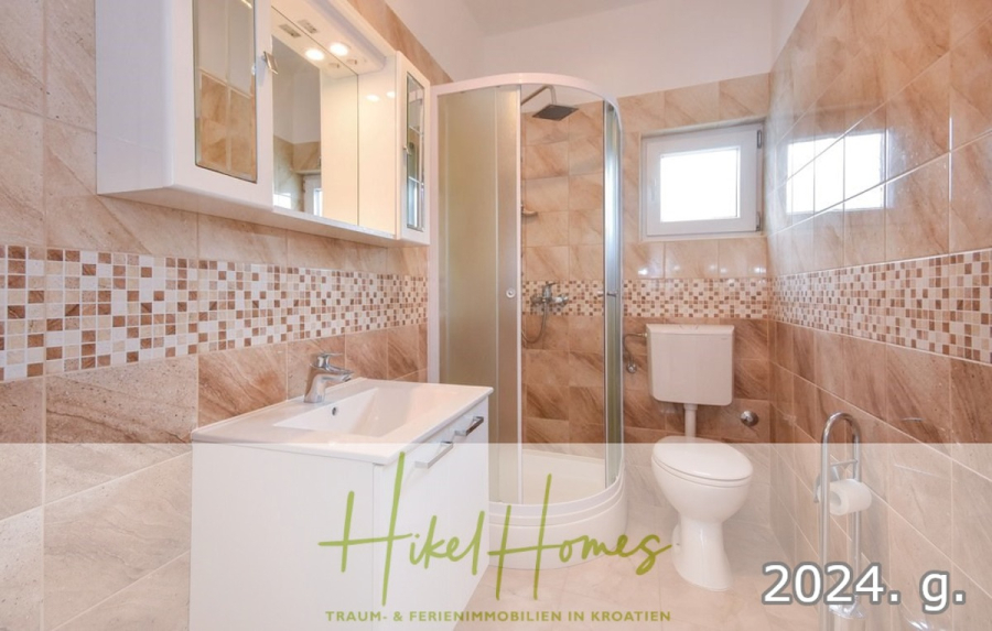 1. Reihe dirkt Meer mit 5 SZ, Pool, Jacuzzi, … auf der Insel Ciovo (bei Trogir mit 2 Brücken) - Modernes Badezimmer mit beigen und braunen Fliesen, einer Glasdusche, einem weißen Waschbecken mit Schubladen, einem Wandspiegelschrank, einer Toilette und einem kleinen Fenster. Die Textüberlagerung lautet "Hikel Homes" und "2024. g. (44929)