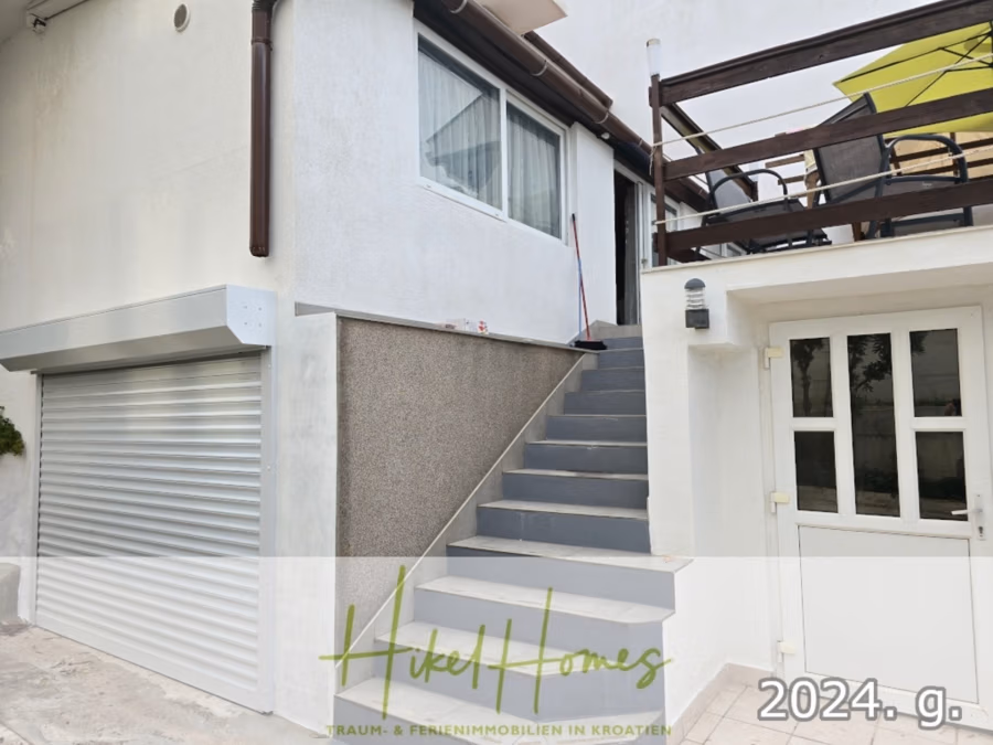 1. Reihe dirkt Meer mit 5 SZ, Pool, Jacuzzi, … auf der Insel Ciovo (bei Trogir mit 2 Brücken) - Außenansicht eines modernen weißen Hauses mit einer Treppe, die zu einer Terrasse führt. Ein großes Fenster und eine Garage mit Rollladen sind sichtbar. Es gibt eine Terrasse mit einer Bank und einem Sonnenschirm. Die Textüberlagerung lautet „Hikel Homes“ und „2024. g.“. (42946)