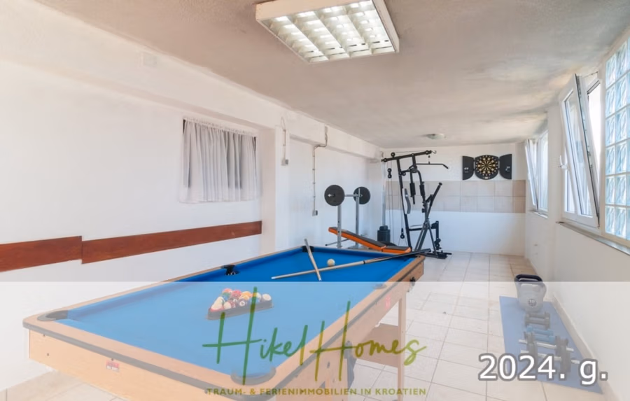 1. Reihe dirkt Meer mit 5 SZ, Pool, Jacuzzi, … auf der Insel Ciovo (bei Trogir mit 2 Brücken) - Ein Freizeitraum mit einem blauen Billardtisch und darauf angeordneten Bällen. Im Hintergrund stehen Fitnessgeräte und eine Dartscheibe. Der Raum hat weiße Wände und ein kleines Fenster mit Vorhängen. Die Texteinblendung lautet „Hikel Homes“ und „2024. g.“. (42943)