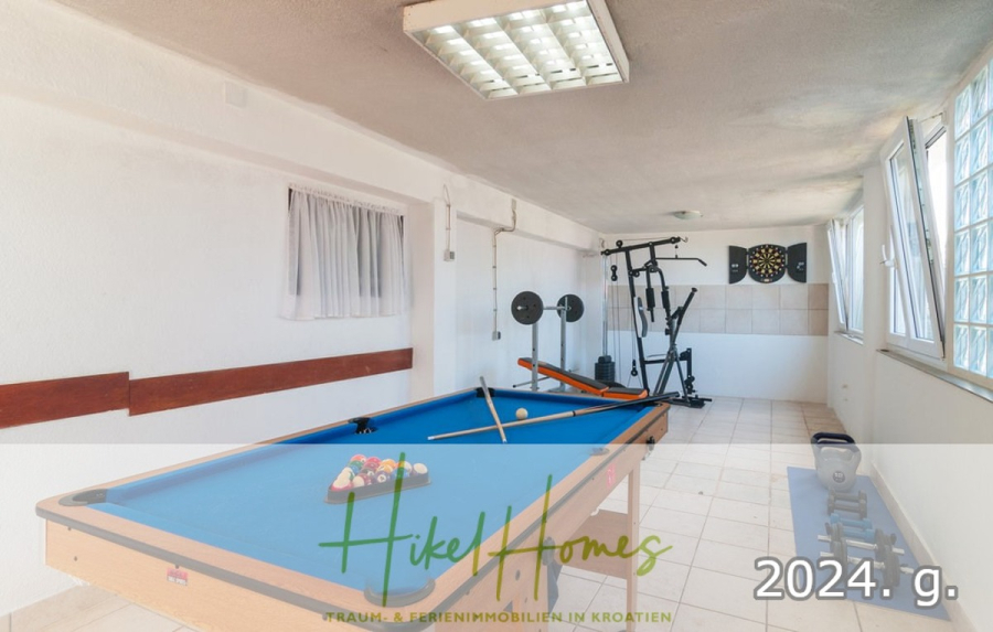 1. Reihe dirkt Meer mit 5 SZ, Pool, Jacuzzi, … auf der Insel Ciovo (bei Trogir mit 2 Brücken) - Ein helles, gefliestes Spielzimmer mit einem blauen Billardtisch, Geräten zum Gewichtheben, einer Dartscheibe an der Wand und großen Fenstern, die natürliches Licht hereinlassen. Das Logo von Hikel Homes und "2024. g." sind am unteren Rand eingeblendet. (44933)