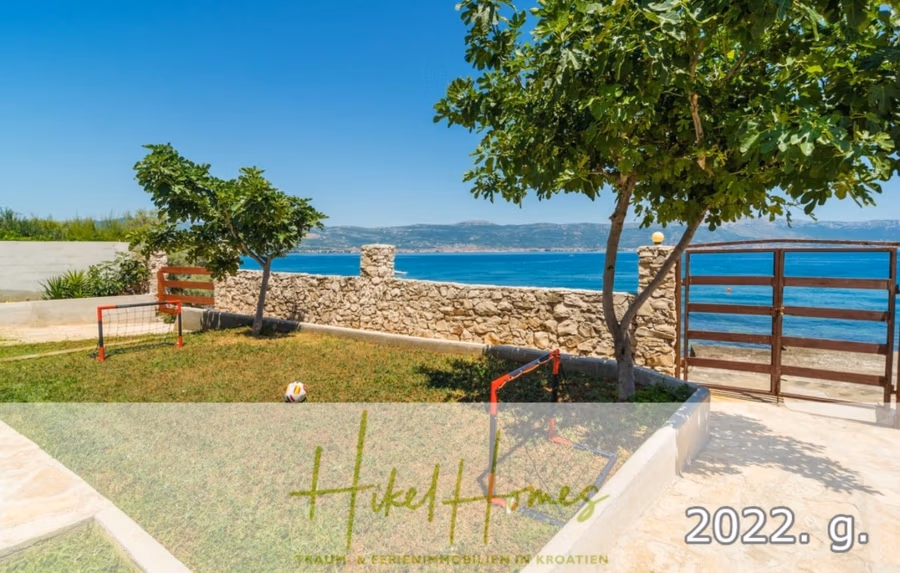 1. Reihe dirkt Meer mit 5 SZ, Pool, Jacuzzi, … auf der Insel Ciovo (bei Trogir mit 2 Brücken) - Üppiger, grüner Garten mit kleinen Fußballtoren, umgeben von einer Steinmauer und mit Blick auf das Meer. Einige Bäume spenden Schatten. Der Himmel ist klar und sonnig. Texteinblendungen zeigen „Hikel Homes“ und „2022“. (42922)