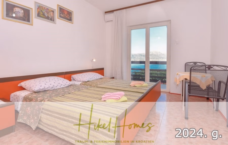1. Reihe dirkt Meer mit 5 SZ, Pool, Jacuzzi, … auf der Insel Ciovo (bei Trogir mit 2 Brücken) - Ein gemütliches Schlafzimmer mit Doppelbett, gemusterter Bettwäsche und gefalteten Handtüchern. Es gibt einen Balkon mit Meerblick, einen kleinen Esstisch mit Stühlen und Wandkunst. Textüberlagerung: „Hikel Homes – Traum- & Ferienimmobilien in Kroatien 2024 g.“. (42932)