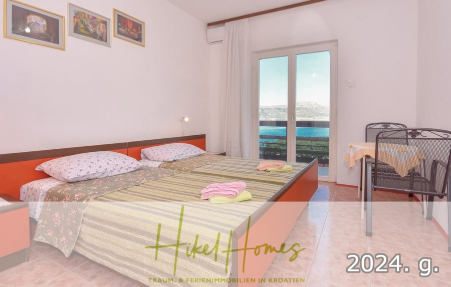 1. Reihe dirkt Meer mit 5 SZ, Pool, Jacuzzi, … auf der Insel Ciovo (bei Trogir mit 2 Brücken) - Ein helles Schlafzimmer mit einem Doppelbett, farbenfroher Bettwäsche, gerahmten Bildern an der Wand, einem kleinen Tisch mit zwei Stühlen und einem Balkon mit Panoramablick. Der Text auf dem Bild lautet "Hikel Homes" und "2024. g.". (44921)