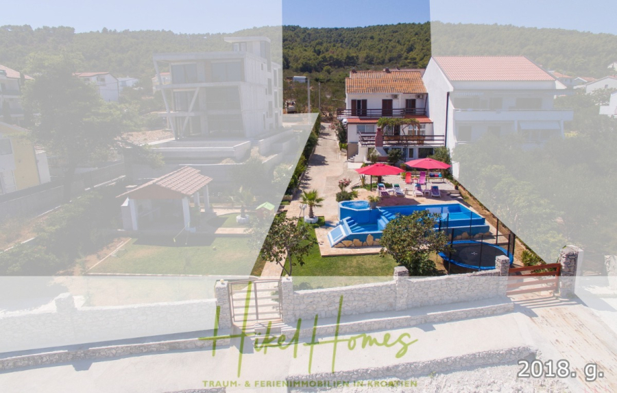 1. Reihe dirkt Meer mit 5 SZ, Pool, Jacuzzi, … auf der Insel Ciovo (bei Trogir mit 2 Brücken) - Luftaufnahme eines Grundstücks mit einem blauen Swimmingpool, Sonnenliegen, Palmen und einem Haus im Grünen. Im Hintergrund sind benachbarte Gebäude und ein Wald am Hang zu sehen. Das Logo "Hitzel Homes" überlagert das Bild. (44938)