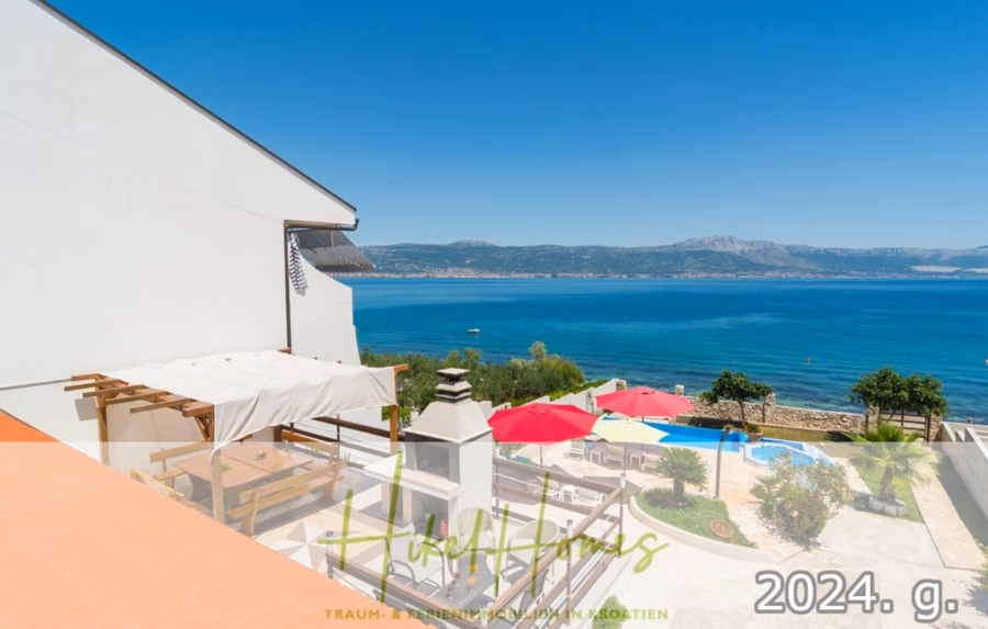 1. Reihe dirkt Meer mit 5 SZ, Pool, Jacuzzi, … auf der Insel Ciovo (bei Trogir mit 2 Brücken) - Ein Küstenblick von einem Balkon: strahlend blaues Meer, Berge in der Ferne und darunter eine Terrasse mit Pergola, roten Sonnenschirmen und grünen Pflanzen. Links ist ein modernes weißes Gebäude zu sehen. Text überlagert das Bild. (42930)