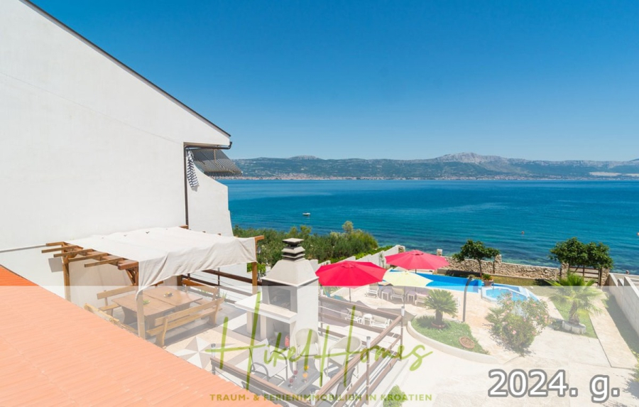 1. Reihe dirkt Meer mit 5 SZ, Pool, Jacuzzi, … auf der Insel Ciovo (bei Trogir mit 2 Brücken) - Blick auf eine sonnige Terrasse mit Tischen, roten Sonnenschirmen und einem weißen Gebäude mit Blick auf ein blaues Meer und ferne Berge. Der Text "Herbsttage 2024. g." erscheint am unteren Rand des Bildes. (44919)