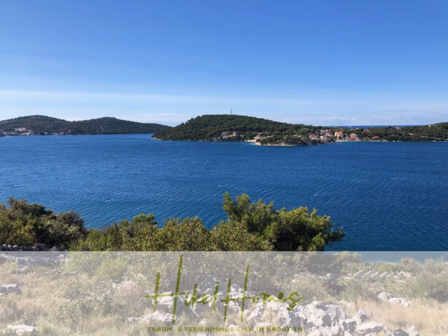 Luxus Villa Zlato – sehr ruhige Wohngegend – eigener Meerzugang, Boje, Pool, Lift und 391m² WFL - Ein malerischer Blick auf eine ruhige blaue Bucht mit üppig grünen Hügeln im Hintergrund unter einem klaren blauen Himmel. Entlang der Küste stehen vereinzelt Gebäude. Im Vordergrund sind Sträucher und Felsen zu sehen. (41818)