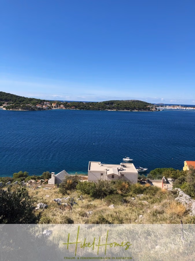 Luxus Villa Zlato – sehr ruhige Wohngegend – eigener Meerzugang, Boje, Pool, Lift und 391m² WFL - Blick auf eine ruhige Küste mit blauem Wasser, grasbewachsenen Hügeln und verstreuten Häusern. Boote säumen den Wasserweg, und in der Ferne ist unter einem klaren blauen Himmel eine bewaldete Insel zu sehen. Unten sind ein Logo und ein Text teilweise sichtbar. (41813)