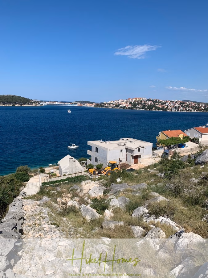 Luxus Villa Zlato – sehr ruhige Wohngegend – eigener Meerzugang, Boje, Pool, Lift und 391m² WFL - Küstenlandschaft mit felsigem Vordergrund und Blick auf das blaue Meer. Mehrere Gebäude, darunter ein weißes Haus, befinden sich in Ufernähe. Ein kleines Boot liegt auf dem Wasser. Am gegenüberliegenden Ufer sind Hügel und weitere Häuser unter einem klaren blauen Himmel zu sehen. (41808)