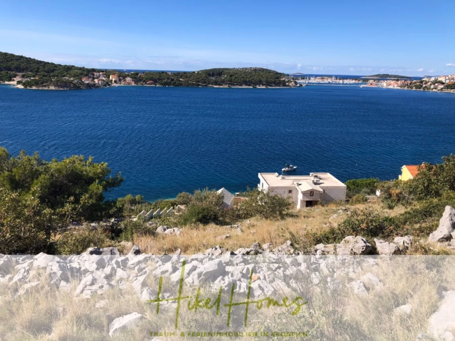 Luxus Villa Zlato – sehr ruhige Wohngegend – eigener Meerzugang, Boje, Pool, Lift und 391m² WFL - Ein malerischer Küstenblick zeigt eine ruhige blaue Bucht, umgeben von hügeligen grünen Inseln und einigen Häusern. Im Vordergrund felsiges Gelände und spärliche Vegetation. Der Himmel ist klar und sonnig. Unten steht „Hikel Homes“. (41817)