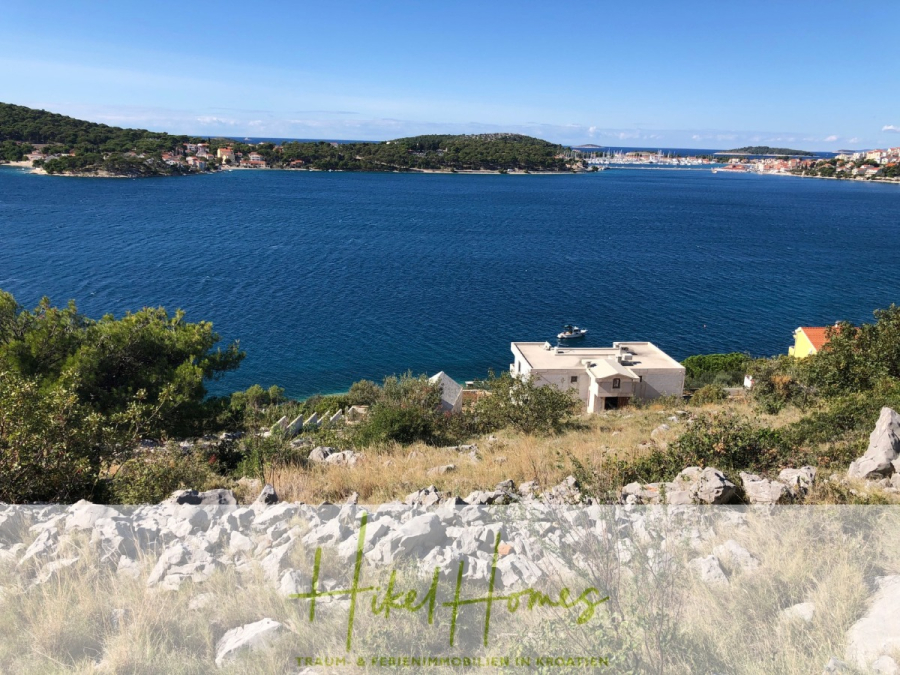 Luxus Villa Zlato – sehr ruhige Wohngegend – eigener Meerzugang, Boje, Pool, Lift und 391m² WFL - Blick auf eine ruhige blaue Bucht, umgeben von grünen Hügeln und kleinen Gebäuden, mit einem felsigen Vordergrund und einem klaren Himmel darüber. Die Worte "Hikel Homes" und ein Slogan erscheinen am unteren Rand des Bildes. (45100)