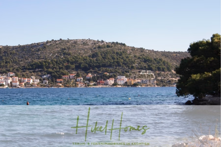 Luxus Villa Zlato – sehr ruhige Wohngegend – eigener Meerzugang, Boje, Pool, Lift und 391m² WFL - Blick auf eine malerische Küstenregion mit klarem, blauem Wasser, einer hügeligen, baumbestandenen Landschaft und einer kleinen Stadt mit farbenfrohen Gebäuden im Hintergrund. Ein Baum rahmt die rechte Seite der Szene teilweise ein. Unten steht der Text „HikelHomes“. (41806)