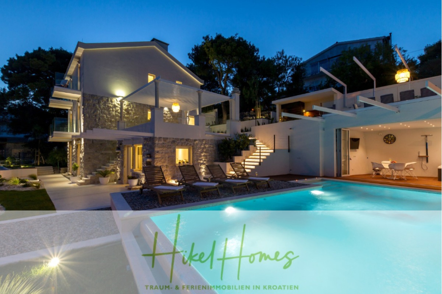 Sehr großzügige Meerblick Villa mit 300m² auf 800m² Grundstück, 4 Garagen, Pool, Terrassen - Moderne Villa mit Außenpool und Liegestühlen, die in der Abenddämmerung beleuchtet werden. Das Gebäude verfügt über Balkone, große Fenster und umliegende Grünflächen. Der Text darüber lautet "Hikel Homes Traum- & Ferienimmobilien in Kroatien". (44904)