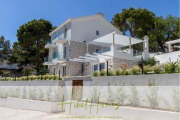 Modernes Haus in Weiß und Stein mit großen Fenstern und Balkonen, umgeben von Grün und an einem sonnigen Hang gelegen. Ein Glasgeländer säumt die Terrasse, und am unteren Rand des Bildes erscheint das Branding "Hikel Homes". - Sehr großzügige Meerblick Villa mit 300m² auf 800m² Grundstück, 4 Garagen, Pool, Terrassen