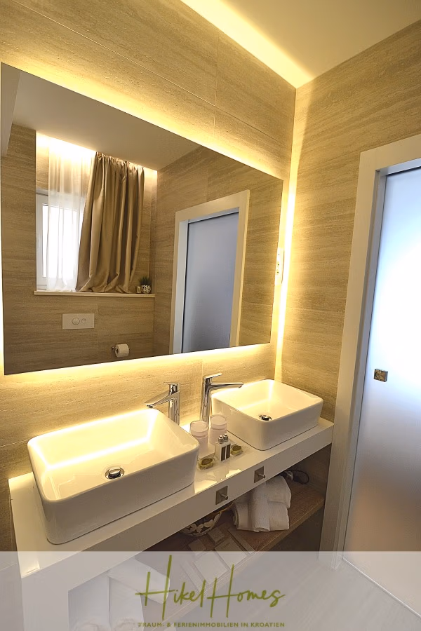 Sehr großzügige Meerblick Villa mit 300m² auf 800m² Grundstück, 4 Garagen, Pool, Terrassen - Modernes Badezimmer mit zwei weißen Waschbecken auf einer Arbeitsplatte, einem großen Spiegel mit Hintergrundbeleuchtung und einem Fenster mit transparenten Vorhängen. Die Wände haben eine helle Holzstruktur und eine teilweise sichtbare Milchglastür. (43280)