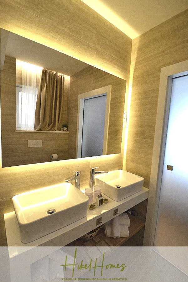 Sehr großzügige Meerblick Villa mit 300m² auf 800m² Grundstück, 4 Garagen, Pool, Terrassen - Modernes Badezimmer mit zwei weißen Waschbecken, silbernen Armaturen, einem großen hinterleuchteten Spiegel, beigen Fliesenwänden, einer Milchglastür und ordentlich angeordneten Toilettenartikeln auf dem Tresen. Die Handtücher werden in den Regalen darunter aufbewahrt. (44893)