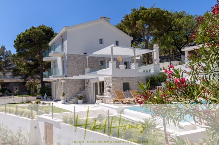 Sehr großzügige Meerblick Villa mit 300m² auf 800m² Grundstück, 4 Garagen, Pool, Terrassen - Eine moderne Steinvilla mit großen Glasbalkonen, umgeben von Bäumen und einem Garten. Im Vordergrund ein Pool mit Sonnenliegen, darüber ein strahlend blauer Himmel. Text: „Hike Homes – Traum- & Ferienimmobilien in Kroatien.“. (43260)