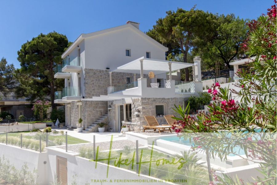 Sehr großzügige Meerblick Villa mit 300m² auf 800m² Grundstück, 4 Garagen, Pool, Terrassen - Moderne Steinvilla mit Glasbalkonen, umgeben von üppigem Grün und einer Poolterrasse mit Liegestühlen unter einem klaren blauen Himmel. Die Umgebung wirkt friedlich und einladend, ideal für einen Urlaubsaufenthalt. (44872)