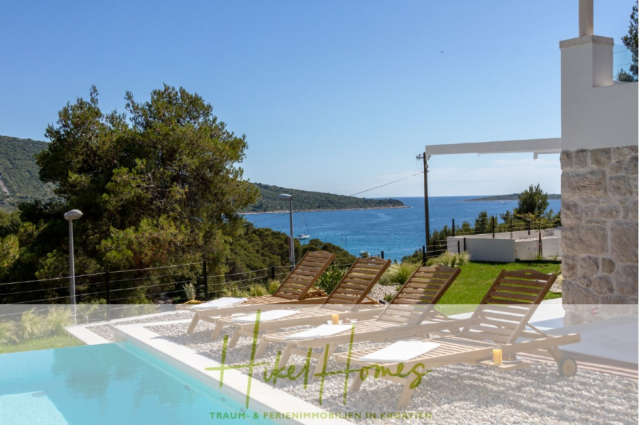 Sehr großzügige Meerblick Villa mit 300m² auf 800m² Grundstück, 4 Garagen, Pool, Terrassen - Vier hölzerne Liegestühle sitzen an einem Pool mit Blick auf das Meer, mit grünen Hügeln und Bäumen im Hintergrund unter einem klaren blauen Himmel; unten steht "Hideaways Homes". (44878)