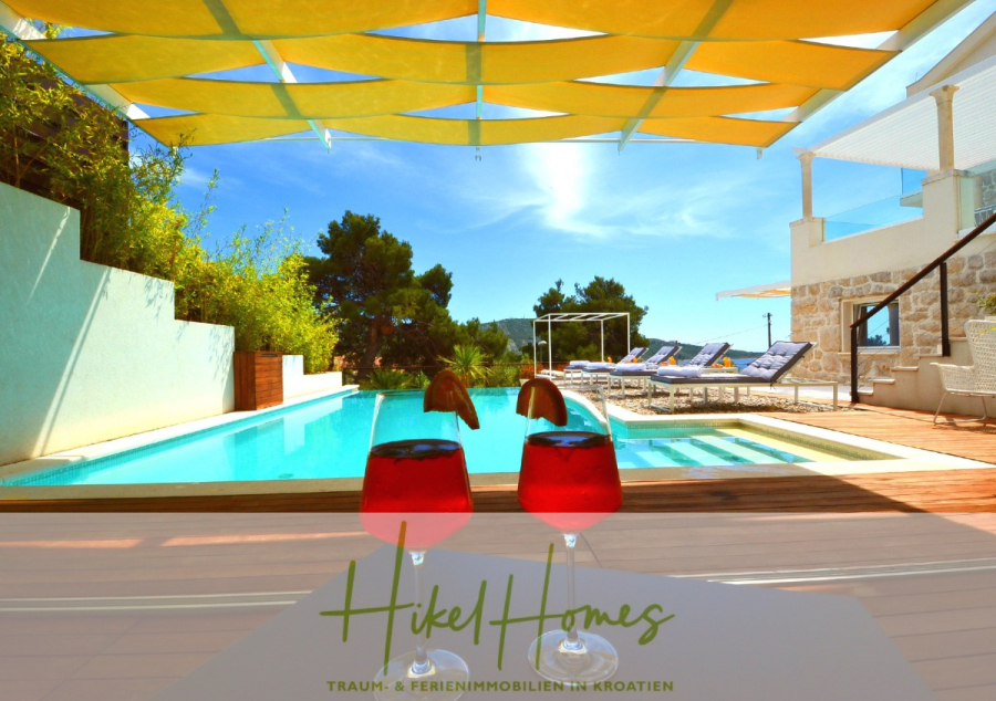 Sehr großzügige Meerblick Villa mit 300m² auf 800m² Grundstück, 4 Garagen, Pool, Terrassen - Zwei Gläser mit roten Getränken stehen auf einem Tisch an einem Pool mit Liegestühlen unter einem gelben Sonnenschirm. Im Hintergrund sind Bäume und moderne weiße Gebäude zu sehen. Das Logo "Hikel Homes" ist am unteren Rand zu sehen. (44877)