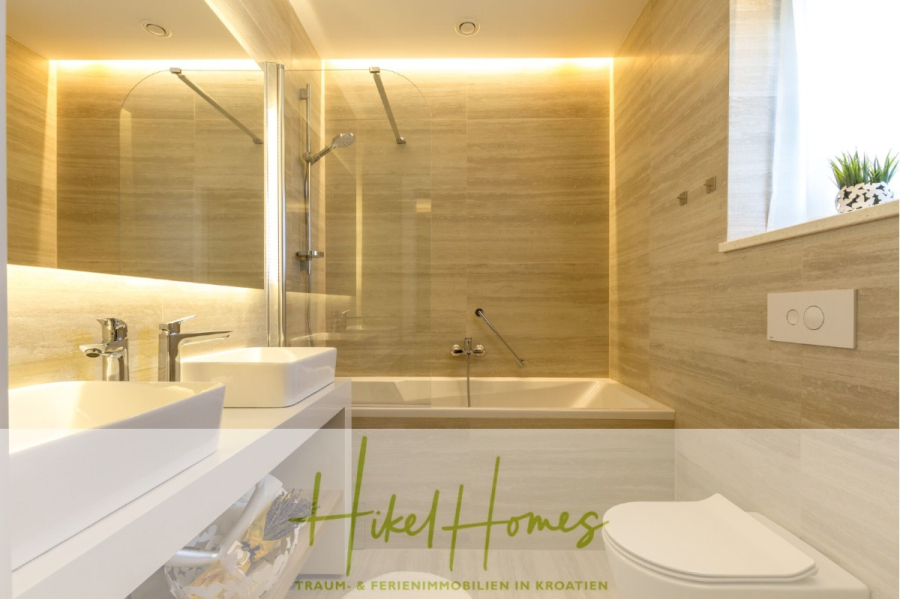 Sehr großzügige Meerblick Villa mit 300m² auf 800m² Grundstück, 4 Garagen, Pool, Terrassen - Modernes Badezimmer mit großem Spiegel, Badewanne mit Dusche, wandmontierter Toilette, weißem Waschbecken, beigen Steinfliesen und einer kleinen Pflanze neben einem Milchglasfenster. Die warme Beleuchtung schafft eine gemütliche Atmosphäre. Der Text lautet: Hikel Homes. (44896)