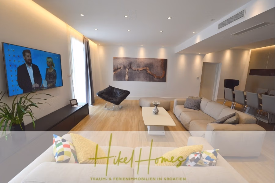 Sehr großzügige Meerblick Villa mit 300m² auf 800m² Grundstück, 4 Garagen, Pool, Terrassen - Modernes Wohnzimmer mit beigefarbenem Sofa, bunten Kissen und einem abstrakten Gemälde an der Wand. Auf einem Flachbildfernseher sind zwei Personen im Gespräch zu sehen. Der Raum verfügt über Holzboden, Einbauleuchten und einen Glasesstisch mit Stühlen. Die Textblende trägt den Text „Hikel Homes“. (43271)