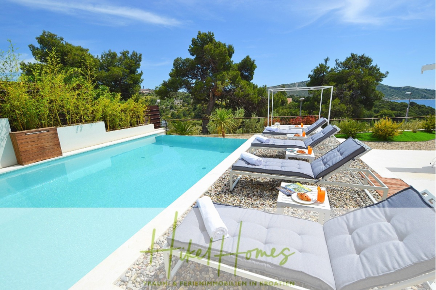 Sehr großzügige Meerblick Villa mit 300m² auf 800m² Grundstück, 4 Garagen, Pool, Terrassen - Moderner Poolbereich mit gepolsterten Liegestühlen, kleinen Tischen mit Getränken und Obst und üppiger Bepflanzung. Der türkisfarbene Pool blickt auf eine malerische Landschaft mit Hügeln und Bäumen unter einem klaren blauen Himmel. (44876)