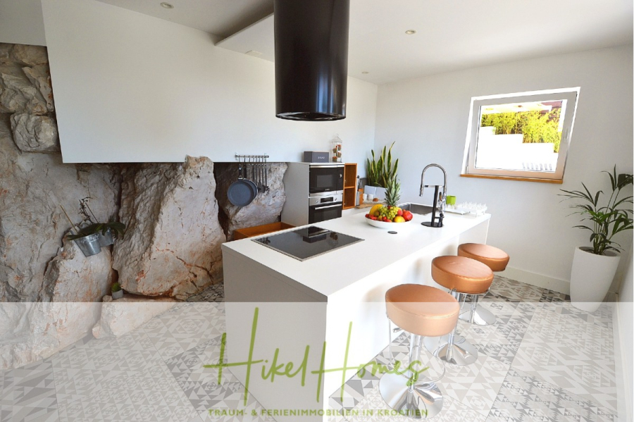 Sehr großzügige Meerblick Villa mit 300m² auf 800m² Grundstück, 4 Garagen, Pool, Terrassen - Moderne weiße Küche mit zentraler Insel, drei braunen Hockern, Einbaugeräten und einer Wand aus Naturstein; durch ein Fenster neben einer Topfpflanze fällt Sonnenlicht ein. (44888)