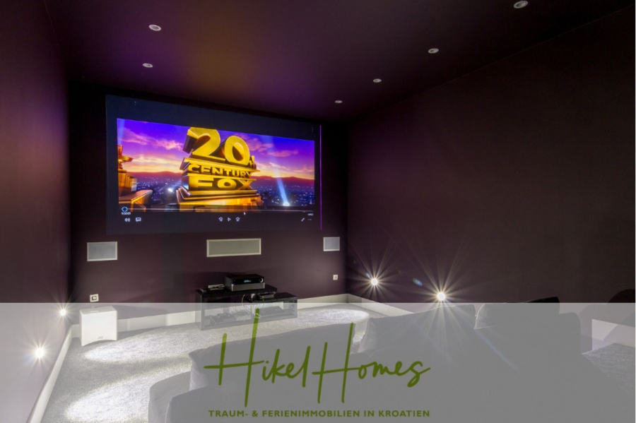 Sehr großzügige Meerblick Villa mit 300m² auf 800m² Grundstück, 4 Garagen, Pool, Terrassen - Ein moderner Heimkinoraum mit dunklen Wänden, einem großen Bildschirm mit dem Logo von 20th Century Fox, Surround-Lautsprechern und silbernen Sitzmöbeln. Das Bild hat eine transparente Überlagerung mit dem Text "Hikel Homes" und einem Slogan auf Deutsch. (44900)