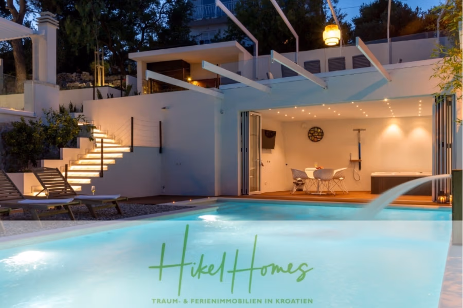 Sehr großzügige Meerblick Villa mit 300m² auf 800m² Grundstück, 4 Garagen, Pool, Terrassen - Modernes Haus mit Essbereich im Freien und Pool. Die Terrasse ist mit einem gedeckten Tisch und einem dekorativen runden Wandelement ausgestattet. Beleuchtete Stufen führen zum Eingang, umgeben von Bäumen und stimmungsvoller Beleuchtung. Auf dem Bild ist der Text „Hikel Homes“ zu sehen. (43289)