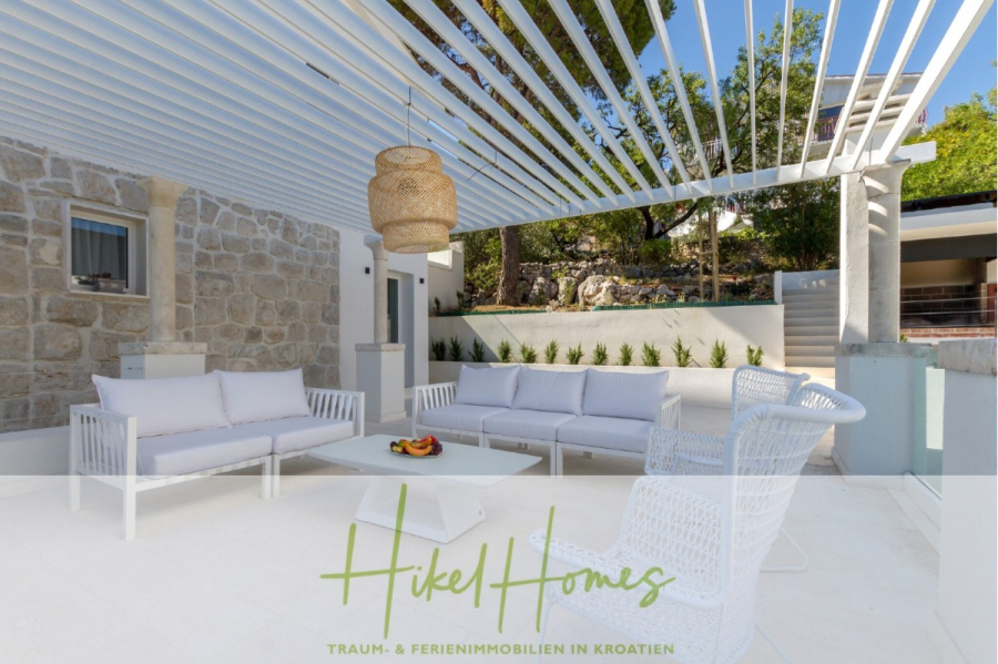 Sehr großzügige Meerblick Villa mit 300m² auf 800m² Grundstück, 4 Garagen, Pool, Terrassen - Moderne Außenterrasse mit weißen Möbeln, einer Steinmauer und einer stilvollen Pergola. Eine geflochtene Pendelleuchte hängt über einem zentralen Couchtisch mit Obst. Das üppige Grün und das sonnige Wetter schaffen eine entspannte Atmosphäre. Der Text lautet: "Hikel Homes. (44882)