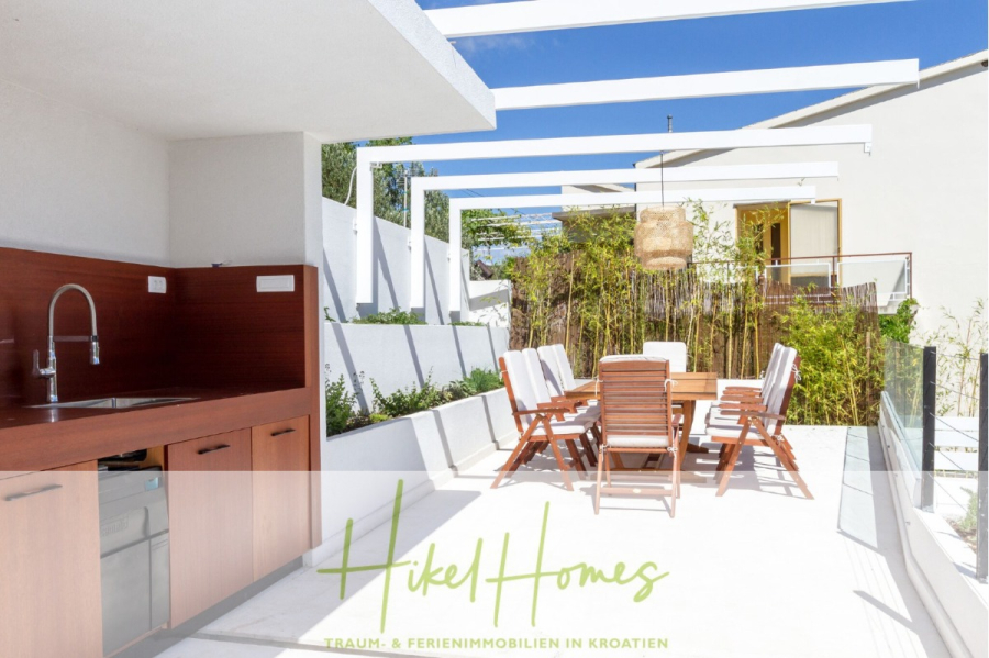 Sehr großzügige Meerblick Villa mit 300m² auf 800m² Grundstück, 4 Garagen, Pool, Terrassen - Eine moderne Außenterrasse mit einem Esstisch aus Holz, Stühlen und einer eingebauten Spüle unter weißen Pergolabalken. Grüne Pflanzen säumen die weißen Wände, und das Sonnenlicht erhellt den klaren, minimalistischen Raum. (44881)