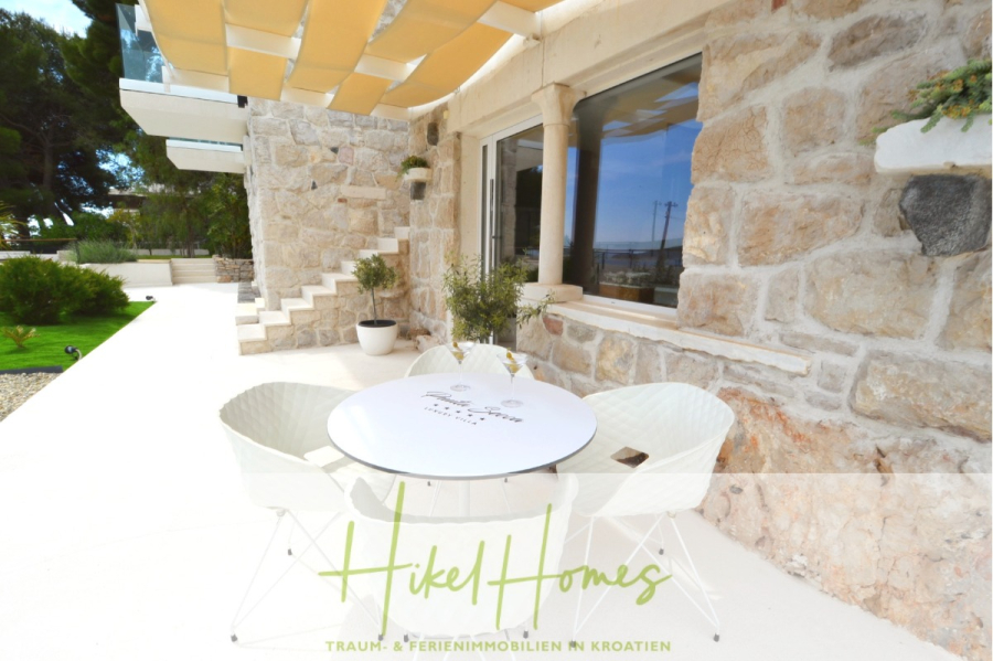 Sehr großzügige Meerblick Villa mit 300m² auf 800m² Grundstück, 4 Garagen, Pool, Terrassen - Eine Steinterrasse mit einem runden weißen Tisch und zwei weißen Stühlen vor einem Steinhaus. Grünpflanzen schmücken den Raum, und große Fenster überblicken die Gegend. Der Text lautet: "Hikel Homes - Traum- & Ferienimmobilien in Kroatien. (44880)