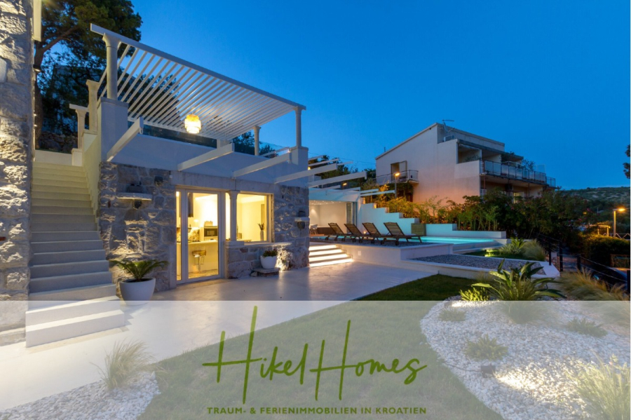 Sehr großzügige Meerblick Villa mit 300m² auf 800m² Grundstück, 4 Garagen, Pool, Terrassen - Moderne Villa mit Steinmauern, Außenbeleuchtung, einem Pool mit Liegestühlen und angelegten Gärten in der Abenddämmerung. Das Logo "Hikel Homes" und ein deutscher Text über Ferienimmobilien in Kroatien erscheinen am unteren Rand. (44901)