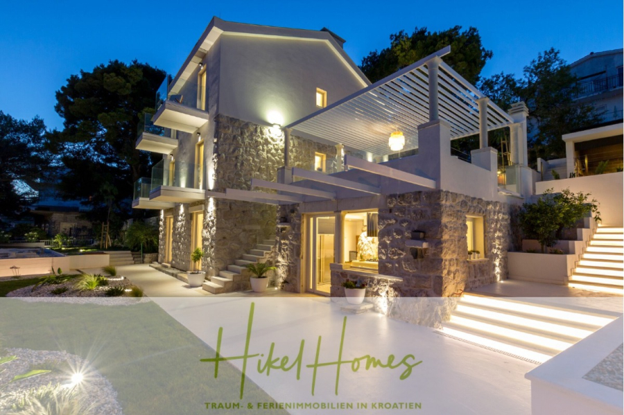Sehr großzügige Meerblick Villa mit 300m² auf 800m² Grundstück, 4 Garagen, Pool, Terrassen - Moderne Steinvilla mit großen Fenstern, Terrassen und Außenbeleuchtung, umgeben von Bäumen in der Abenddämmerung. "Hikel Homes: Traum- & Ferienimmobilien in Kroatien" steht am unteren Rand. (44906)