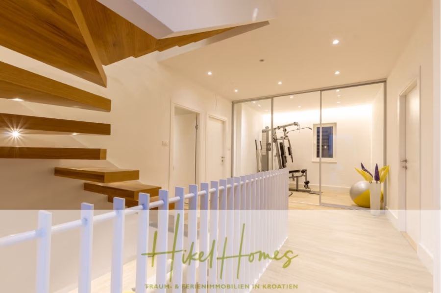 Sehr großzügige Meerblick Villa mit 300m² auf 800m² Grundstück, 4 Garagen, Pool, Terrassen - Modernes Interieur mit freitragender Holztreppe links, verglastem Fitnessbereich mit Trainingsgeräten und hellem Holzboden. Weißes Lattengeländer und helle, eingelassene Deckenleuchten runden den klaren, modernen Look ab. (43285)