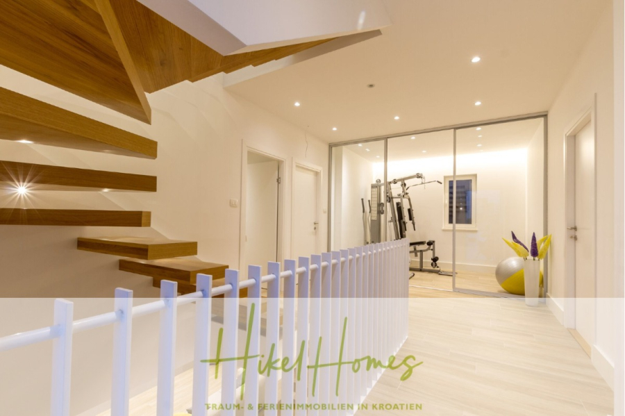 Sehr großzügige Meerblick Villa mit 300m² auf 800m² Grundstück, 4 Garagen, Pool, Terrassen - Moderner, heller Flur mit schwebender Holztreppe, gläserner Fitnessbereich mit Fitnessgeräten und Gymnastikbällen. Weißes Geländer und schlichtes, minimalistisches Dekor. Text "Hikel Homes" am unteren Rand. (44898)