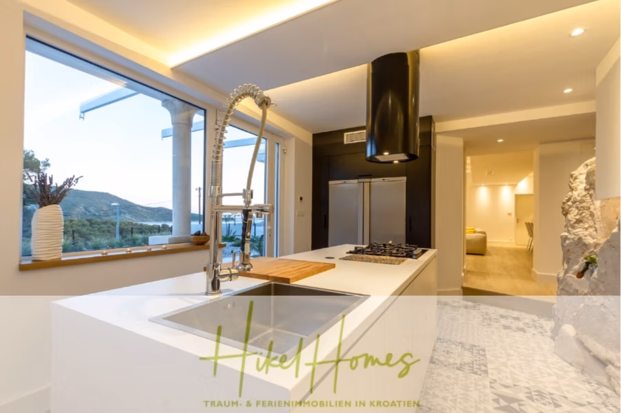 Sehr großzügige Meerblick Villa mit 300m² auf 800m² Grundstück, 4 Garagen, Pool, Terrassen - Moderne Küche mit großen Fenstern und herrlicher Aussicht. Die elegante weiße Kücheninsel verfügt über einen Chromhahn und einen eingebauten Herd. Darüber hängt eine schwarze Dunstabzugshaube. Der Raum geht in einen sanft beleuchteten, minimalistischen Wohnbereich über. (43276)