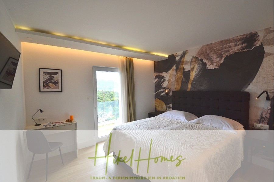 Sehr großzügige Meerblick Villa mit 300m² auf 800m² Grundstück, 4 Garagen, Pool, Terrassen - Modernes Hotelzimmer mit einem Doppelbett, einem abstrakten Wandgemälde, einem Schreibtisch mit Stuhl, einem an der Wand montierten Fernseher und einer Glastür, die auf einen Balkon mit Blick ins Freie führt. Der Raum ist hell und übersichtlich. (44894)