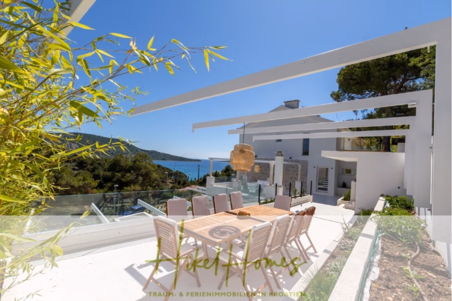 Sehr großzügige Meerblick Villa mit 300m² auf 800m² Grundstück, 4 Garagen, Pool, Terrassen - Dachterrasse mit Esstisch und Stühlen aus Holz mit Blick auf das Meer und die grünen Hügel. Moderne weiße Gebäude umgeben das Gelände, mit klarem blauen Himmel und Blick auf üppiges Grün. Eine Bambuspflanze sorgt für einen Hauch von Natur. (43262)