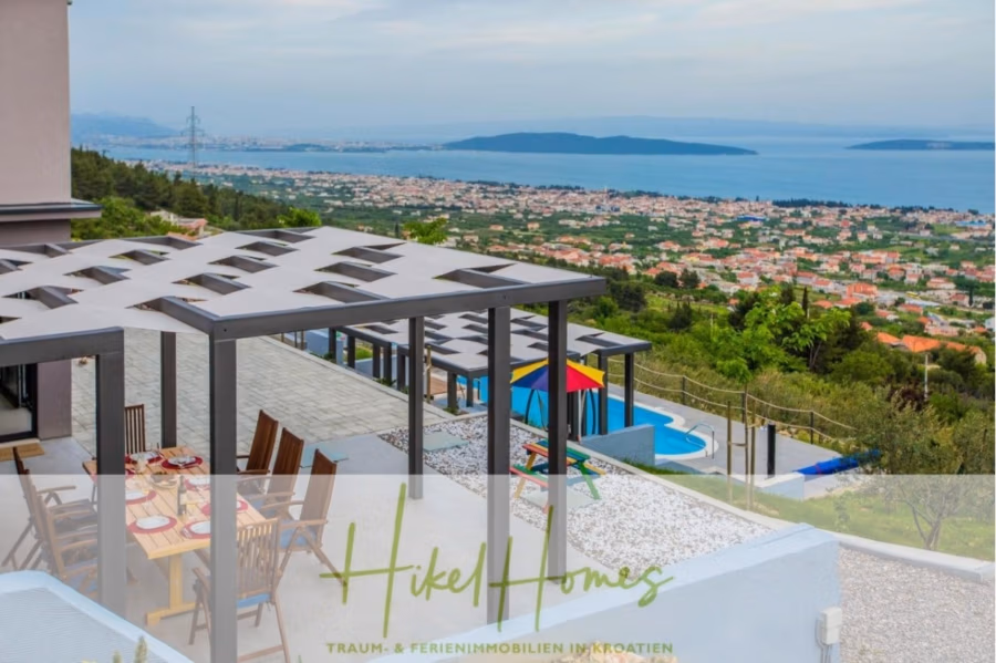 Meerblick auf 2903m² großem Grundstück in wunderbarer Alleinlage am Stadtrand von Split - Die Dachterrasse mit Essbereich bietet Blick auf eine Küstenstadt und das Meer. Im Vordergrund befinden sich eine moderne Pergola und ein Pool mit bunten Sonnenschirmen, umgeben von viel Grün. Im Hintergrund sind Hügel und ein bewölkter Himmel zu sehen. (41453)