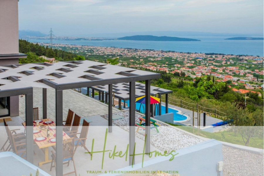 Meerblick auf 2903m² großem Grundstück in wunderbarer Alleinlage am Stadtrand von Split - Eine moderne Terrasse mit Esstischen bietet einen Blick auf einen Pool mit bunten Sonnenschirmen und eine malerische Küstenstadt, umgeben von Grün und Bergen unter einem bewölkten Himmel. Der Text "Hikel Homes" ist am unteren Rand des Bildes zu sehen. (45870)
