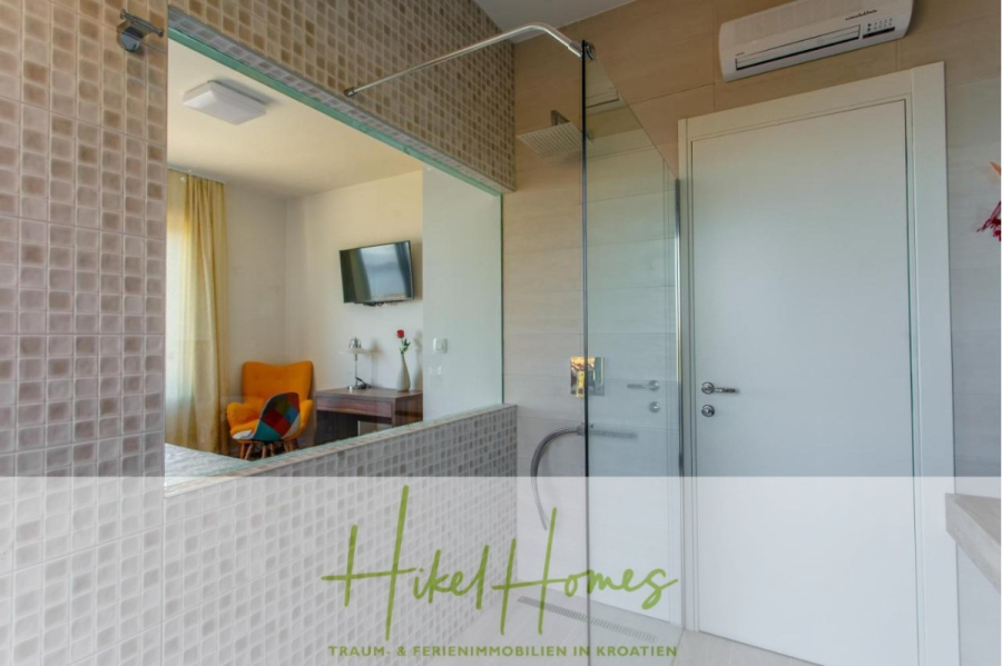 Meerblick auf 2903m² großem Grundstück in wunderbarer Alleinlage am Stadtrand von Split - Modernes Badezimmer mit einer Glasdusche, beige gefliesten Wänden, einem großen Spiegel und einem Blick in ein helles Schlafzimmer mit einem gelben Stuhl, einem Schreibtisch und einem an der Wand befestigten Fernseher. Das Logo "Hikel Homes" erscheint am unteren Rand des Bildes. (45853)