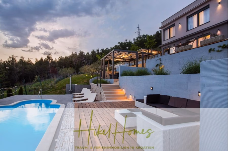 Meerblick auf 2903m² großem Grundstück in wunderbarer Alleinlage am Stadtrand von Split - Moderne Villa am Hang mit Außenpool, der in der Abenddämmerung von warmem Licht erhellt wird. Die Terrasse bietet Sitzgelegenheiten und einen Essbereich, umgeben von Bäumen und sanften Hügeln. Der Himmel ist teilweise bewölkt, was eine ruhige Urlaubsatmosphäre schafft. (41452)